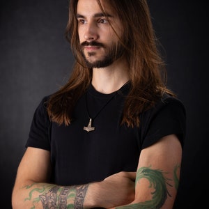 May include: A man with long brown hair and a beard is wearing a black t-shirt with a silver Mjolnir pendant necklace. He has a green dragon tattoo on his right arm and a wolf head and runes tattoo on his left arm.