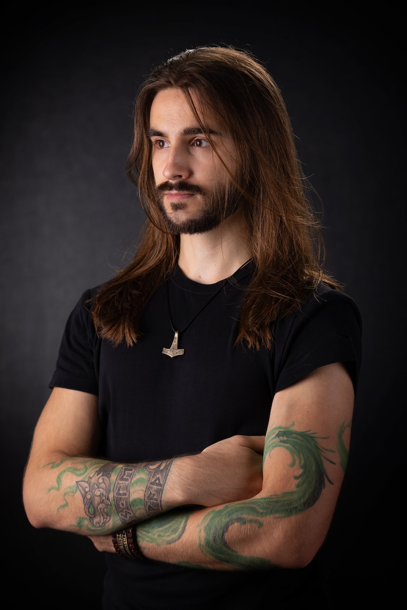 May include: A man with long brown hair and a beard is wearing a black t-shirt with a silver Mjolnir pendant necklace. He has a green dragon tattoo on his right arm and a wolf head and runes tattoo on his left arm.