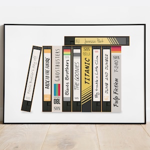 Puede incluir: Una impresión enmarcada de nueve cintas VHS con títulos de películas, incluyendo "Jurassic Park", "Titanic", "Pulp Fiction", "Regreso al futuro", "Cazafantasmas", "Blues Brothers", "Los Goonies", "El día libre de Ferris Bueller" y "Dos tontos muy tontos".