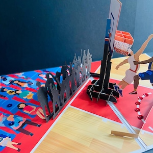 May include: A 3D pop-up basketball card featuring a basketball court scene. The card shows players, a hoop, and a crowd of cheering fans. The court is red and tan, with a blue and red background.