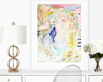May include: Abstract painting in a white frame with a white background, featuring swirls of blue, yellow, pink, and white paint. The painting is on a white desk with a gold lamp and a clear chair.