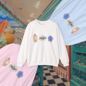May include: A white sweatshirt with three embroidered patches. The patches include a pink and gold candy shop, a green and white logo that says "Honeydukes", and a blue and silver hexagon. Two other sweatshirts, one pink and one blue, are also visible.