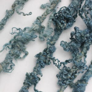 May include: Close-up of several strands of blue-green yarn with a textured, curly appearance. The yarn is arranged diagonally across a white background, showcasing its unique texture and colour variations. Ideal for fibre art projects.