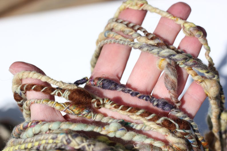 May include: A skein of hand-spun art yarn featuring a textured, uneven ply with a variegated color palette of cream, beige, brown, dark purple, and soft green. The yarn displays varying thicknesses and twists throughout the length of the fiber.