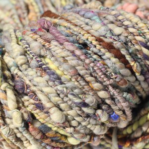 May include: A skein of art yarn featuring a multi-textured, hand-spun appearance with varying thicknesses. The yarn displays a palette of muted earth tones, including beige, brown, soft yellow, dusty purple, and hints of blue, with twisted fibers.