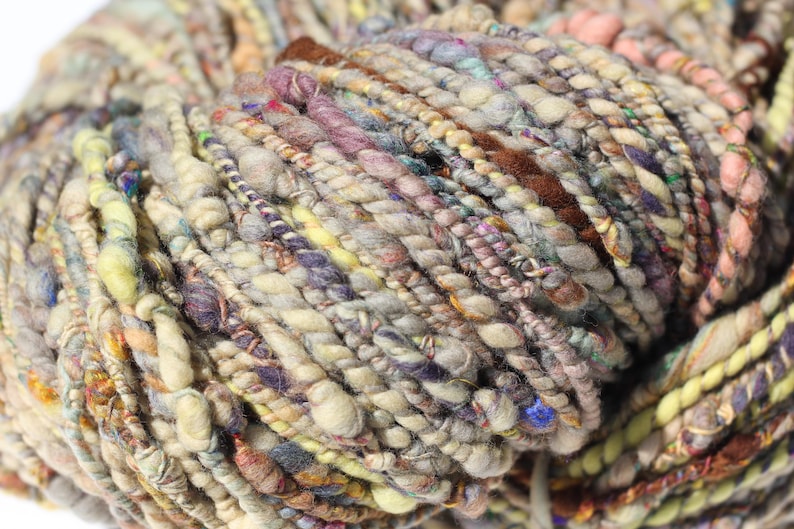 May include: A skein of art yarn featuring a multi-textured, hand-spun appearance with varying thicknesses. The yarn displays a palette of muted earth tones, including beige, brown, soft yellow, dusty purple, and hints of blue, with twisted fibers.