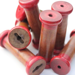 May include: A group of wooden spools with red painted tops. The spools are stacked and arranged in a pile. The spools are made of natural wood and have a smooth, polished finish.