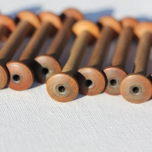 Puede incluir: Una colección de carretes de madera vintage, dispuestos en fila. Los carretes tienen un cuerpo marrón oscuro con un extremo circular marrón claro. Cada carrete tiene un pequeño agujero en el centro. Los carretes están sobre una superficie blanca.
