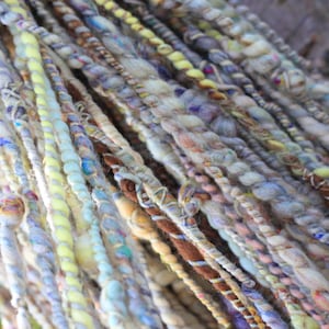 May include: A collection of hand-spun art yarn strands featuring a variety of textures and thicknesses. The fibers display a palette of soft pastel blues, yellows, creams, and earthy browns, twisted together with varying degrees of tension and density.