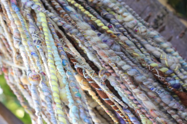 May include: A collection of hand-spun art yarn strands featuring a variety of textures and thicknesses. The fibers display a palette of soft pastel blues, yellows, creams, and earthy browns, twisted together with varying degrees of tension and density.