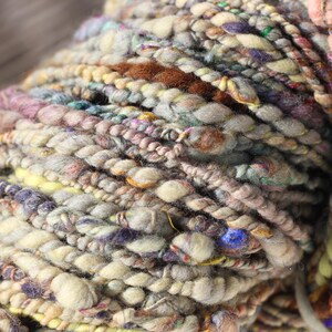 May include: A skein of hand-spun art yarn featuring a thick and thin texture with a multi-colored palette of cream, beige, brown, soft pink, yellow, and deep purple fibers, incorporating twisted strands and small flecks of vibrant blue throughout.