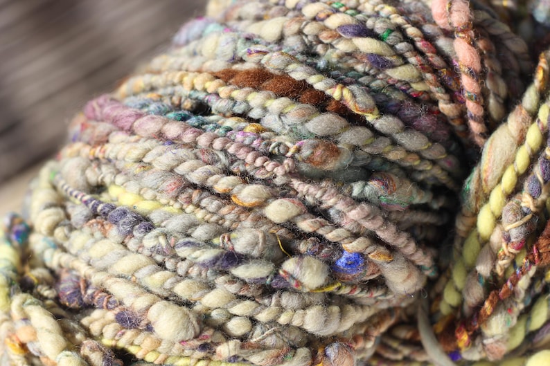 May include: A skein of hand-spun art yarn featuring a thick and thin texture with a multi-colored palette of cream, beige, brown, soft pink, yellow, and deep purple fibers, incorporating twisted strands and small flecks of vibrant blue throughout.