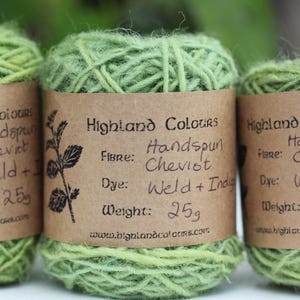 Könnte beinhalten: Drei Stränge grüner handgesponnener Wolle mit braunen Papieretiketten. Die Etiketten tragen die Aufschrift "Highland Colours" und enthalten Angaben zu Faser, Farbstoff und Gewicht von 25g. Das Garn hat eine hellgrüne Farbe.