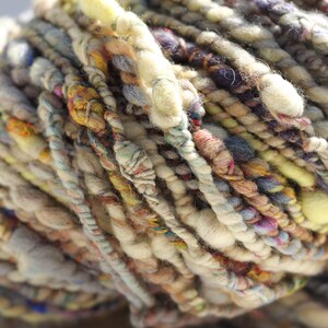 May include: A skein of art yarn featuring a thick and thin texture with a multi-colored palette of cream, beige, brown, yellow, and purple. The yarn consists of various twisted fibers, including slubbed sections and thin strands.