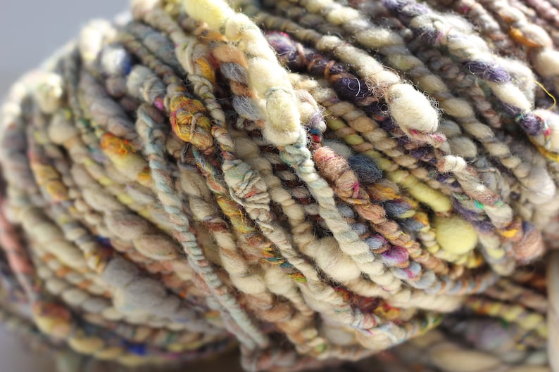 May include: A skein of art yarn featuring a thick and thin texture with a multi-colored palette of cream, beige, brown, yellow, and purple. The yarn consists of various twisted fibers, including slubbed sections and thin strands.