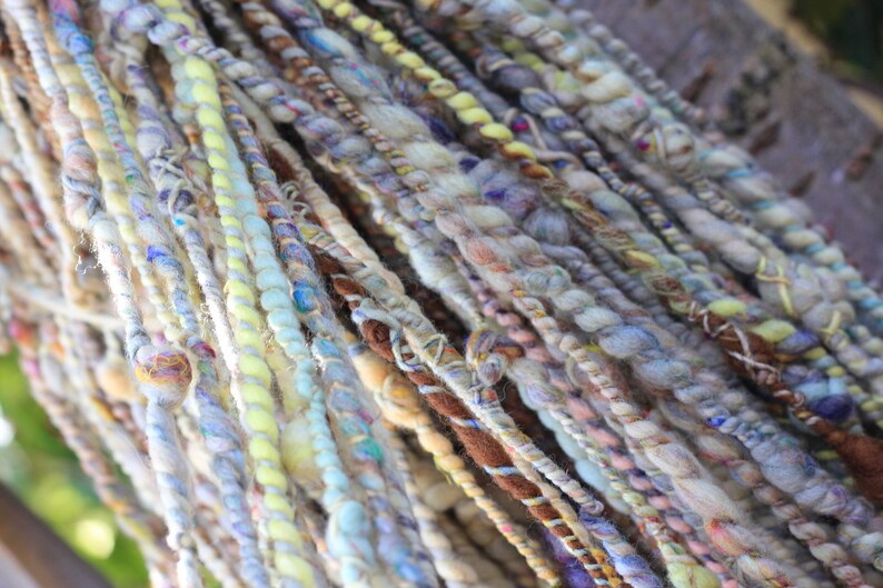 May include: A collection of hand-spun art yarn strands featuring a variety of textures and thicknesses. The fibers display a palette of soft pastel blues, yellows, creams, and earthy browns, twisted together with varying degrees of tension and density.