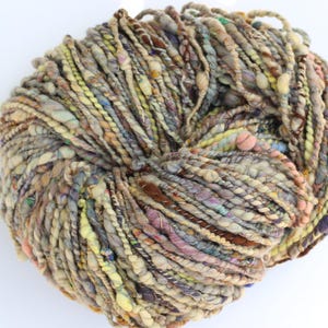 May include: A round skein of hand-spun art yarn featuring a multi-textured, thick-and-thin construction. The yarn displays a variegated color palette of muted greens, browns, soft pinks, blues, and cream, with various fiber types twisted together.
