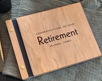 May include: A wooden photo album or guest book with a dark spine and gold-colored metal fasteners. The front cover features the engraved text "CONGRATULATIONS ON YOUR RETIREMENT" and the name "MICHAEL JONES" in a clean, dark sans-serif font.