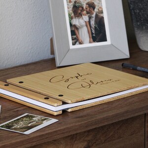 A custom bamboo wedding guest photo album rests on a table beside several photos, ready to be attached to the pages of the book.