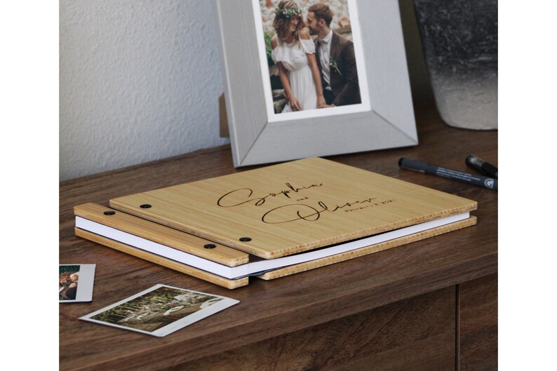 A custom bamboo wedding guest photo album rests on a table beside several photos, ready to be attached to the pages of the book.