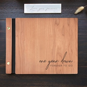A wooden photo album with a black spine and gold accents. The album has the text 'one year down forever to go' engraved on the front.