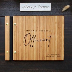 May include: A rectangular bamboo wood binder with a black spine and gold-coloured metal fasteners. The cover is engraved with the text Michael Anderson, Officiant, and Gabby + Jacob September 21, 2023. A separate white sign reads Here's to Forever.