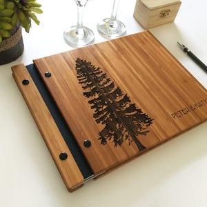 May include: A square bamboo photo album with a laser-engraved pine tree design on the cover. The text "PETER & KATY" is engraved on the lower right side. The album features a dark spine secured with black metal rivets and a natural wood grain.