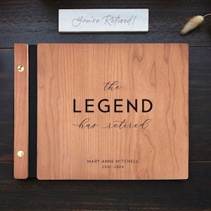 Op de afbeelding: Een rechthoekig houten fotoalbum met een zwarte rug en goudkleurige metalen sluitingen. De kaft bevat de gegraveerde tekst "the LEGEND has retired" samen met de naam "MARY ANNE MITCHELL" en de jaartallen "2001-2024" in een donker lettertype.