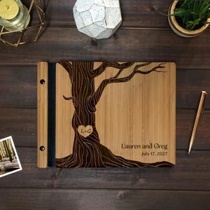 May include: A rectangular bamboo photo album with a dark brown laser-engraved tree design on the cover. The tree trunk features a heart shape with the letters L+G inside. The text Lauren and Greg and the date July 17, 2027 are engraved on the right.