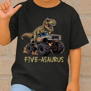 May include: Black t-shirt featuring a green dinosaur driving a monster truck. The text "FIVE-ASARUS" is printed below the graphic. The child is wearing blue jeans and sunglasses.