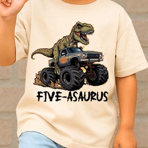 May include: A beige short-sleeved t-shirt featuring a graphic of a green dinosaur riding a monster truck. The text "FIVE-ASASURUS" is printed below the graphic. The child is wearing light blue jeans with ripped knees.