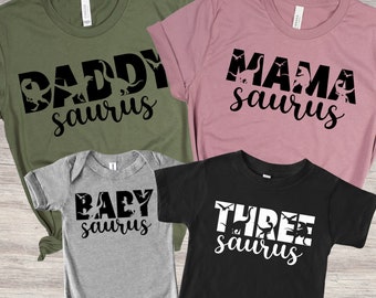 May include: Set of four t-shirts with dinosaur silhouettes and the text "Daddy saurus", "Mama saurus", "Baby saurus", and "Three saurus". The shirts are in green, pink, gray, and black.