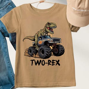 May include: Tan short-sleeved t-shirt featuring a cartoon dinosaur driving a monster truck. The truck has large black tires and the text "TWO-REX" below the image. A tan baseball cap and blue jeans are also visible.
