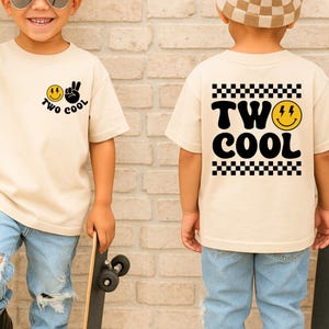 May include: A beige t-shirt with the text "TWO COOL" and a smiley face graphic. The shirt is worn by a child wearing sunglasses and light blue ripped jeans. The child is holding a skateboard.