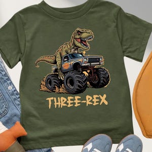 May include: Olive green t-shirt featuring a cartoon dinosaur driving a monster truck. The text "THREE-REX" is printed in a yellow-orange color below the truck. The shirt is displayed with blue jeans, a toy sword, a yellow bag, and blue sneakers.