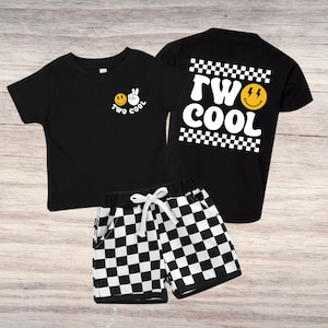 May include: A black two-piece toddler outfit featuring a t-shirt and shorts. The t-shirt has a smiley face and peace sign graphic with the words "TWO COOL". The shorts are black and white checkered. The back of the shirt also says "TWO COOL".