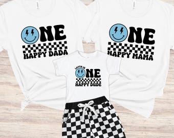 Könnte beinhalten: Zwei weiße T-Shirts mit schwarzen und hellblauen Grafiken mit der Aufschrift ONE HAPPY DADA und ONE HAPPY MAMA, ein kleineres weißes T-Shirt mit der Aufschrift LAYTH IS ONE HAPPY DUDE mit hellblauem Smiley und schwarzem Karomuster sowie schwarz-weiße Karo-Shorts.