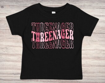 May include: Black toddler t-shirt with the word "THREENAGER" in a layered pink and white design. The text is stacked vertically, with each word slightly offset. The shirt is made of a soft material.