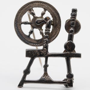 May include: A silver-toned brooch in the shape of a spinning wheel. The brooch features a detailed wheel, spindle, and frame, with a pin on the back for attachment. The design is intricate and likely vintage, suitable for craft enthusiasts or collectors.