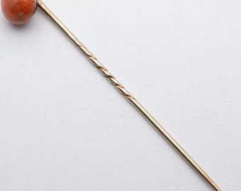 May include: A vintage hat pin with a round, orange-brown stone top. The pin's shaft is gold-toned, with a twisted design near the top. The pin tapers to a sharp point. The background is plain white.