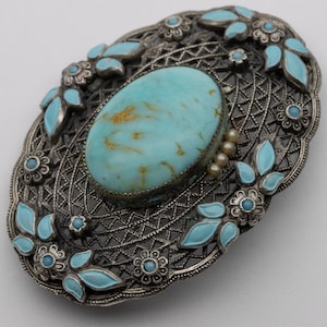 May include: An oval-shaped, antique silver brooch with a large, light blue turquoise stone at the center. The brooch features a filigree design, small turquoise accents, and light blue enamel leaves. A cluster of small pearls is on the side.