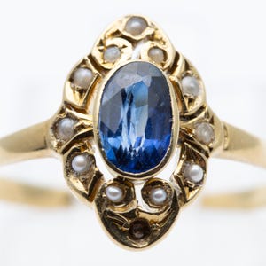 May include: A gold ring featuring an oval, deep blue gemstone at the center, surrounded by small, round pearls. The ring's setting has an ornate, filigree design. The band is a simple, polished gold.