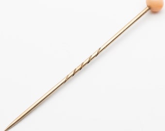 May include: A gold-toned hat pin with a spiral design on the shaft and a peach-colored, spherical head. The pin is pointed at one end.