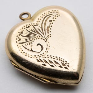 Puede incluir: Un relicario dorado en forma de corazón con un diseño floral grabado en la parte delantera. El relicario tiene un pequeño lazo en la parte superior para una cadena. El diseño grabado presenta un remolino y un patrón de hojas.