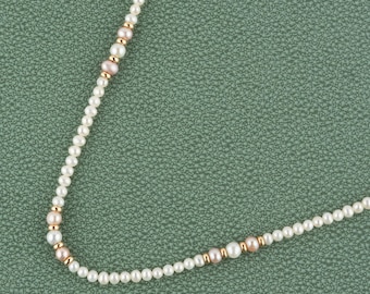 May include: A delicate pearl necklace featuring small, round white pearls interspersed with gold-colored beads and a few pinkish-toned pearls. The necklace is displayed against a textured, muted green background.