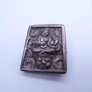 Könnte beinhalten: Rechteckige, silberfarbene Brosche mit geprägtem Design. Die Brosche zeigt eine zentrale Figur mit mehreren Armen und einen detaillierten, verzierten Rand. Das Design umfasst schlangenartige Kreaturen und ein Gesicht am unteren Rand. Die Brosche ist wahrscheinlich Vintage oder antik.