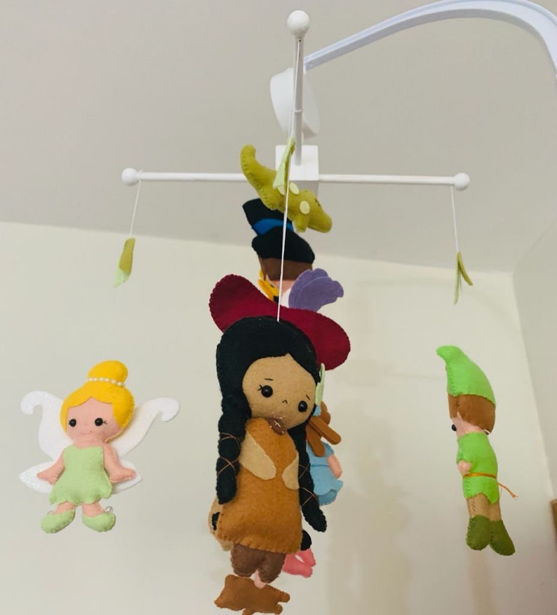 Pu&ograve; includere: Un mobile bianco per neonati con personaggi in feltro appesi. I personaggi includono Peter Pan, Trilly, Pocahontas e un alligatore verde. Il mobile &egrave; progettato per la cameretta di un bambino.