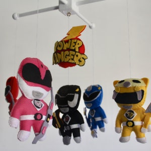 May include: A baby mobile featuring five felt figures of the Power Rangers. The figures are pink, black, blue, yellow, and red. The mobile also includes a felt sign that reads "Power Rangers" with a lightning bolt.