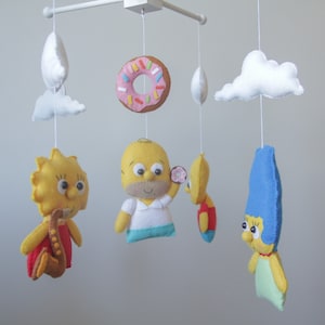 Op de afbeelding: Een babymobiel met een gele donut, witte wolken en viltfiguren van de Simpsons. De figuren zijn Homer, Marge, Lisa en Bart.