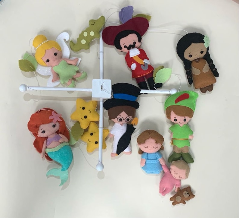 Pu&ograve; includere: Un mobile bianco con personaggi di feltro appesi. I personaggi includono Trilly, Peter Pan, Capitan Uncino, una sirena, una donna nativa americana, un personaggio di Alice nel Paese delle Meraviglie, un bambino con un orsacchiotto e due stelle gialle.